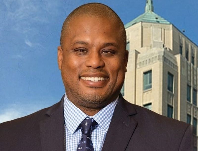 A head shot of a Black man with a shaved head. He is wearing a dark suit jacket, blue checked shirt, and a tie. The top of a light brick building with a pointed light green roof is in the background.