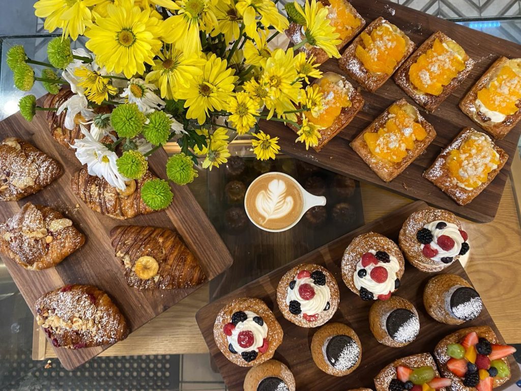 a Birdseye view of a table featuring 3 wooden boards full of croissants and pastries with a latte in the middle. There is a vase of yellow, green, and white flowers in the upper left side of the picture.