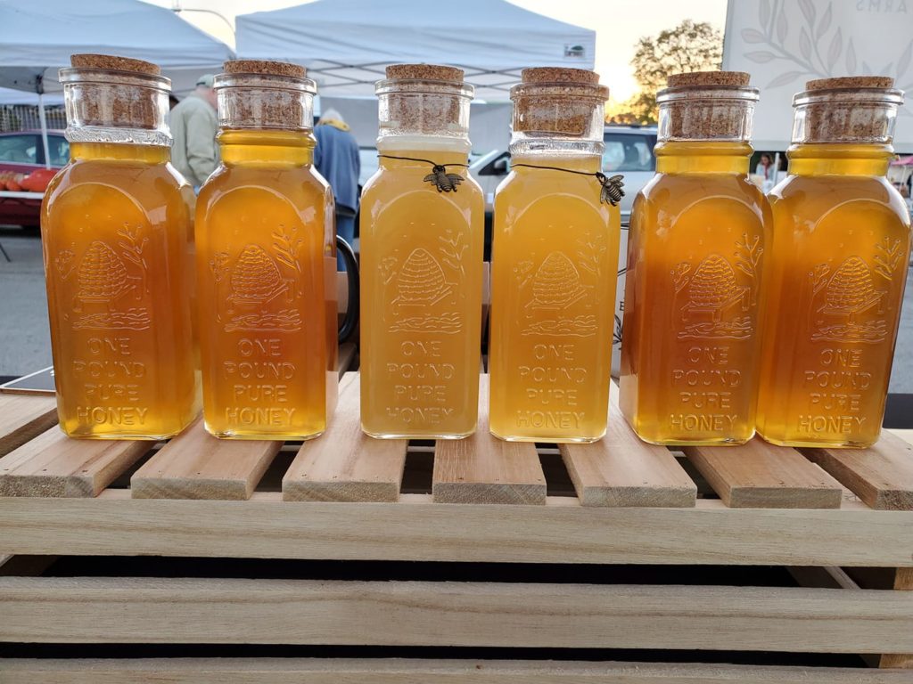 Six jars of honey are displayed on a wood shelf. The bottles are embossed with "one pound pure honey" and stopped with corks. The two bottles in the middle are a lighter but more opaque color than the other four. 