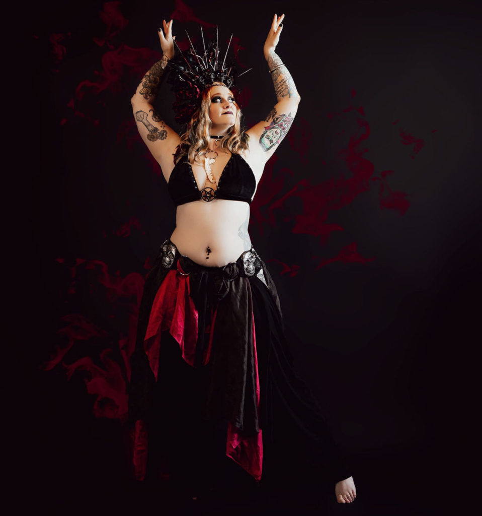 Amanda Garion stands against a red wall, wearing a black bra, and black pants with red fabric hanging down, as well as an elaborate metal crown. Her arms are bent and extended toward the ceiling as she looks up and to the right.
