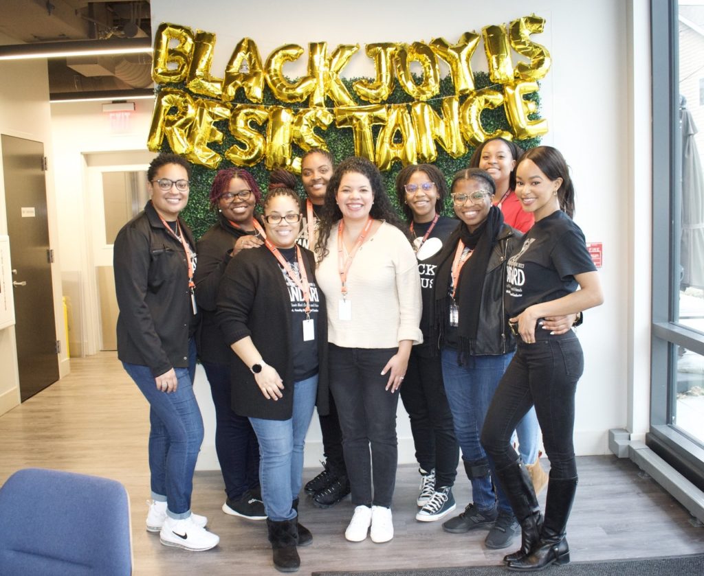 Eight Black women pose in front of a wall hanging that says "Black Joy Is Resistance."They are all smiling at the camera. All are wearing orange lanyards, as if part of a conference. 