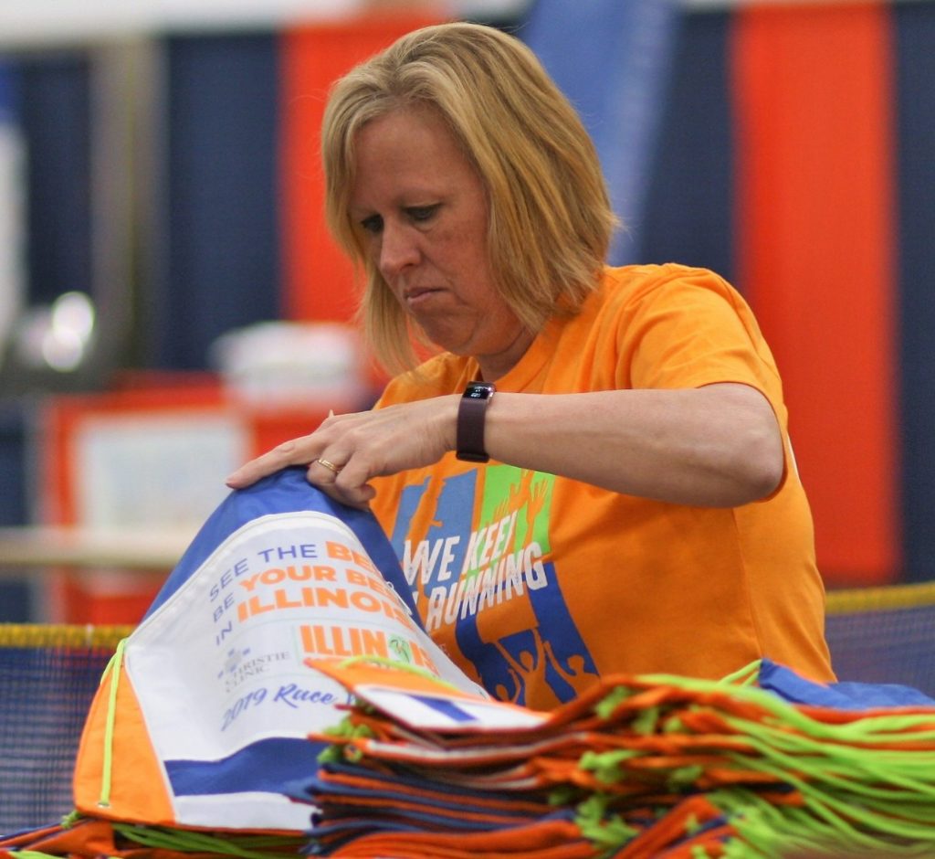 a woman with strawberry blonde hair wears an orange shirt that says, 'we keep running". She is putting something into a bag for race participants. 