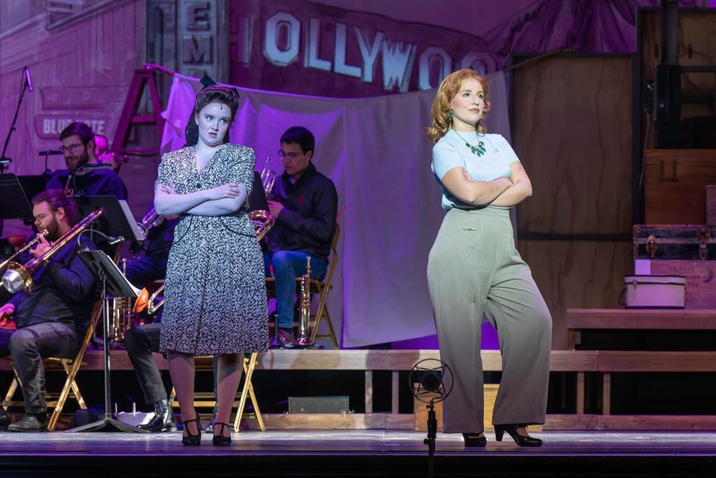 A scene from City of Angels. Two white women are on the stage, both with their arms crossed. On the left, a woman with brown hair is wearing a patterned dress and stares at the back of the head of the woman on the right, a redhead wearing a blouse and trousers. 