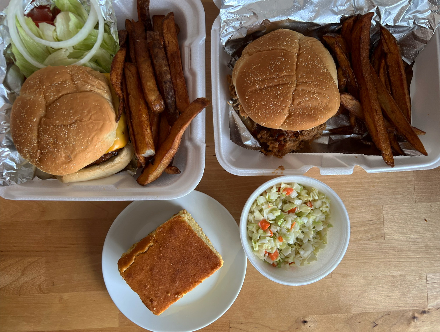 Takeout from Sooie Bros Bar B Que. Two sandwiches with sides of fries are in to-go containers. A slice of cornbread is on a plate next to them, as well as a container of cole slaw.