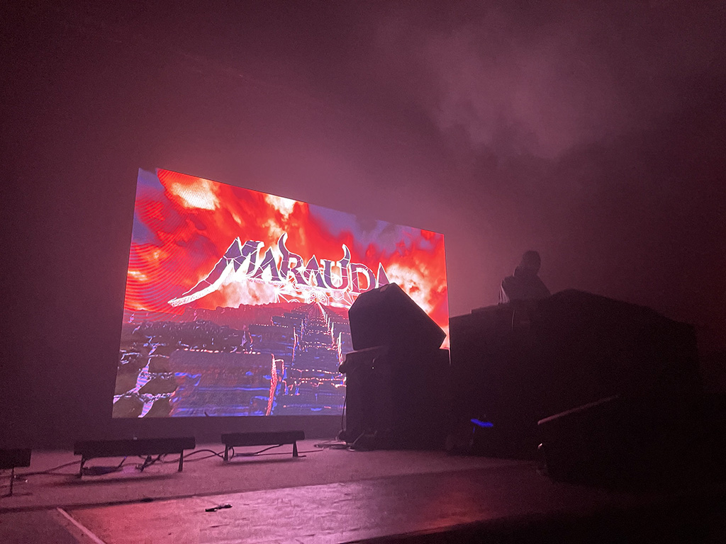 The stage at an EDM show. Marauda is playing his music on the decks onstage, and a video screen with "Marauda" on it is behind him.