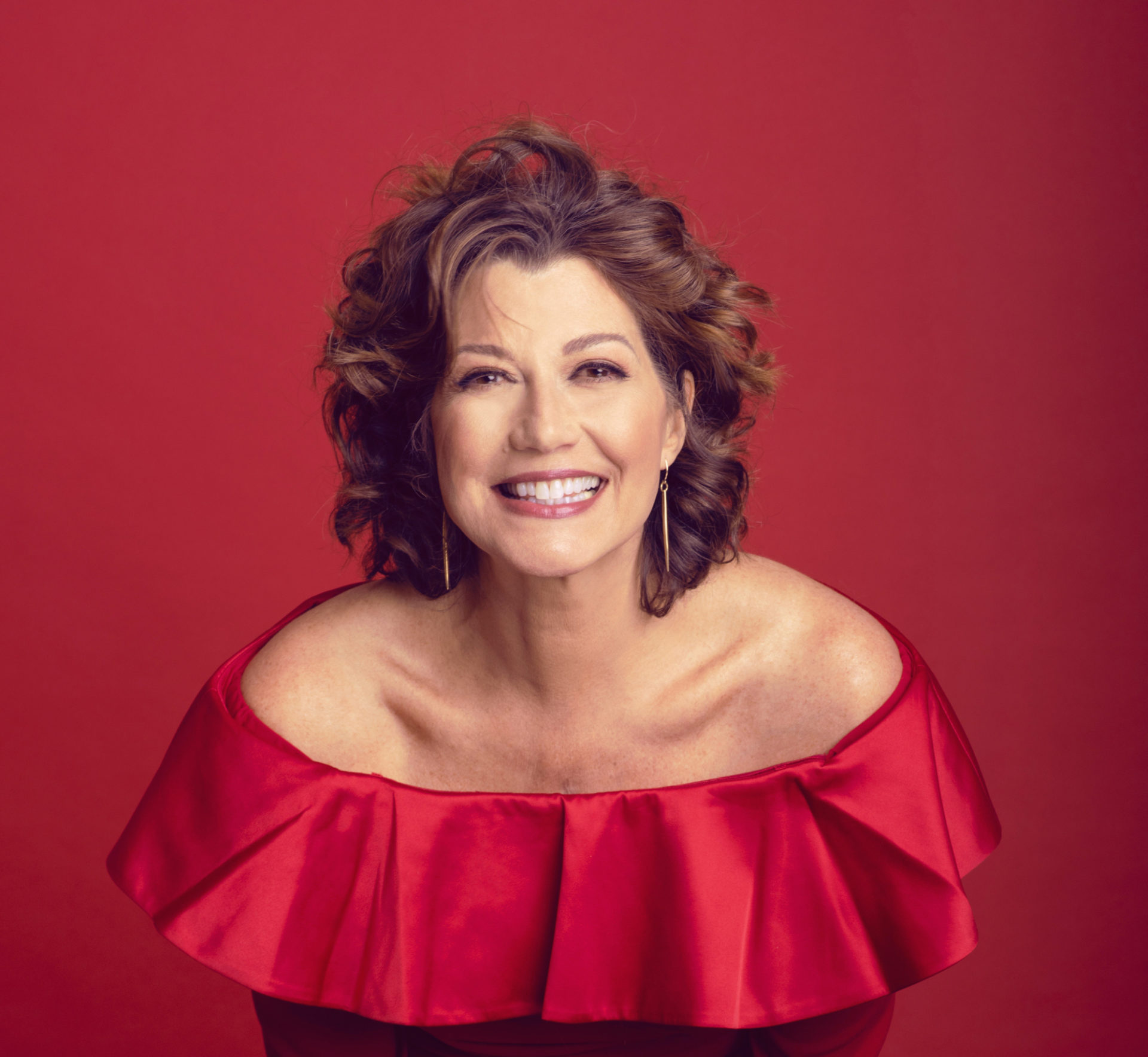 Singer Amy Grant, a white woman, seen in portrait. She is pictured wearing a red, off the shoulder shiny top. Her hair is shoulder length and curly. She is grinning at the camera. The background is red.