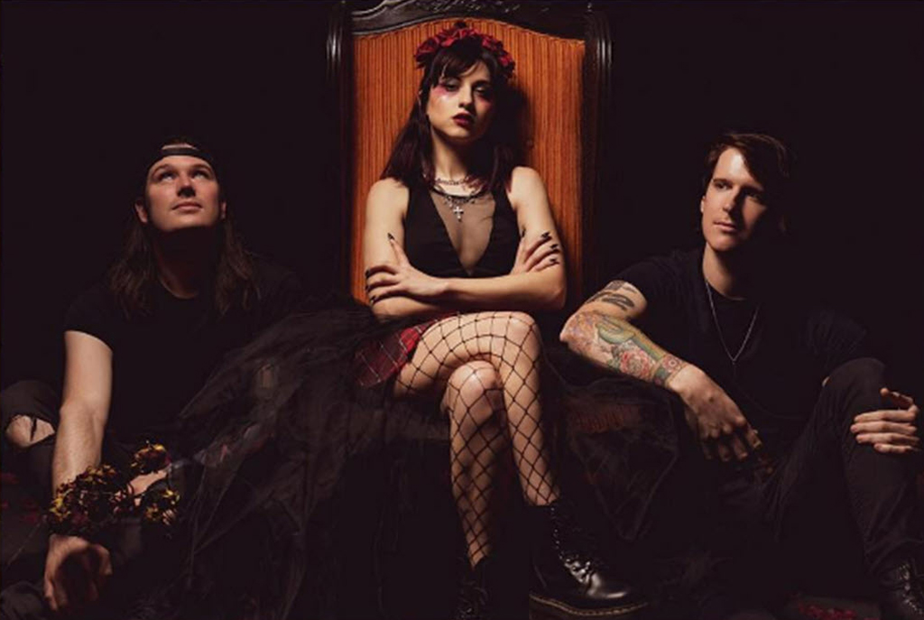 3 members of the band Ashland sitting. Person on left is sitting on the ground looking up. Person in the middle is a woman and is sitting on a chair, crossing her arms and looking at the camera. The person on the right is male and is sitting on the ground looking off to the left.