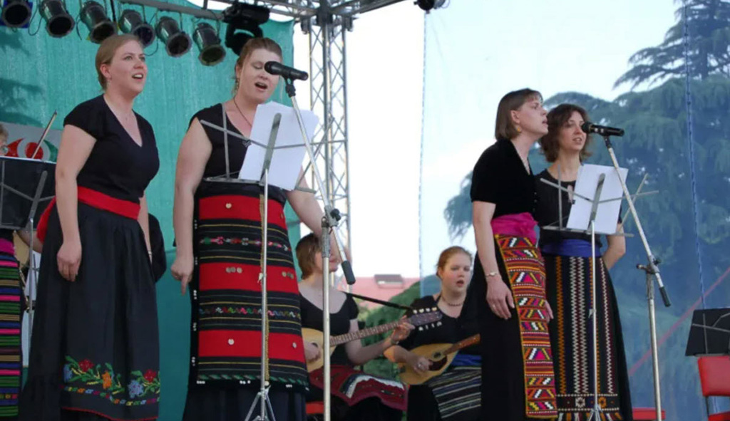 Balkanalia performing on an outdoor stage. There are 4 vocalists up front, and you can see 2 mandolin players in the background.