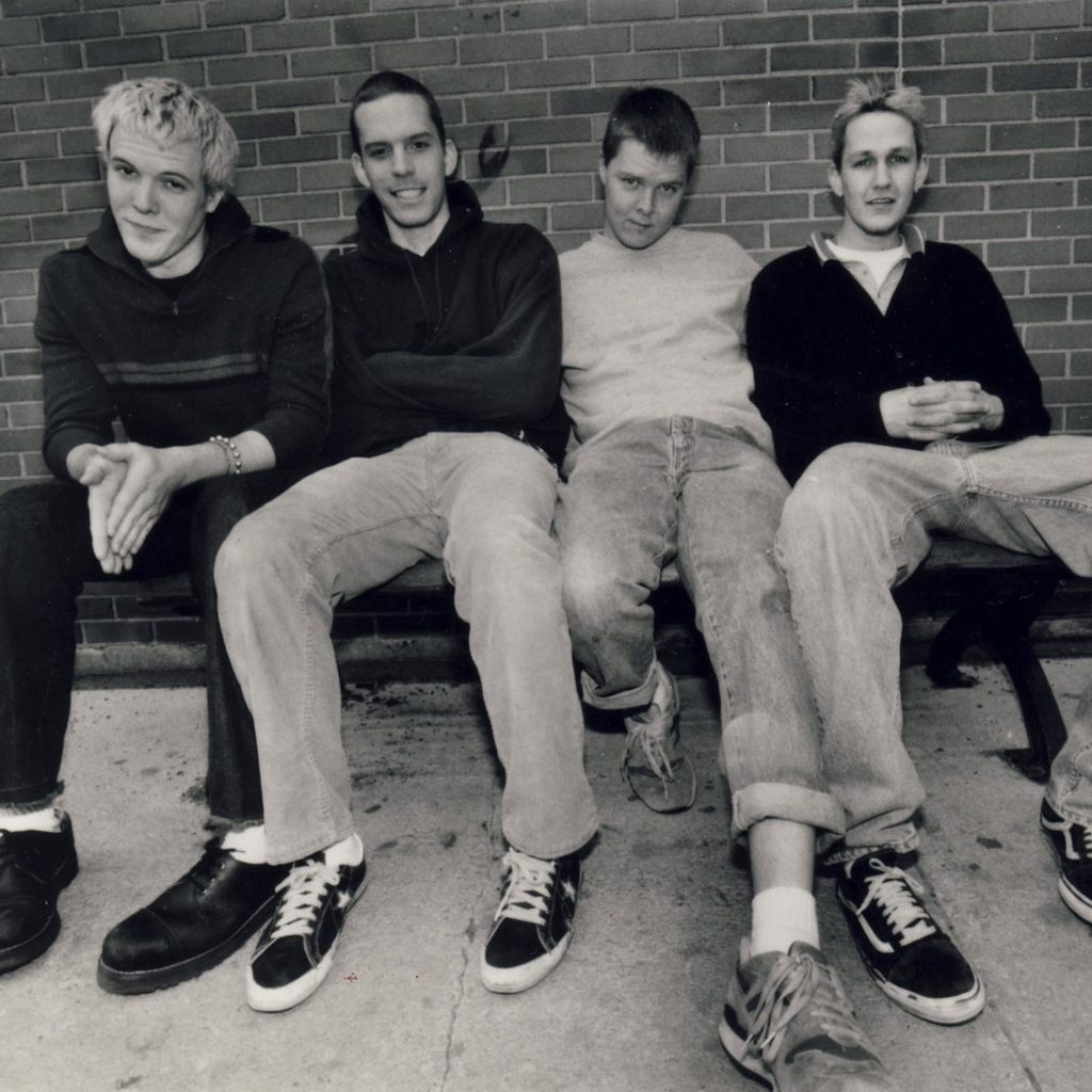 The band Braid in the 1990s. Four young white men sit on a bench. The photo is black and white.