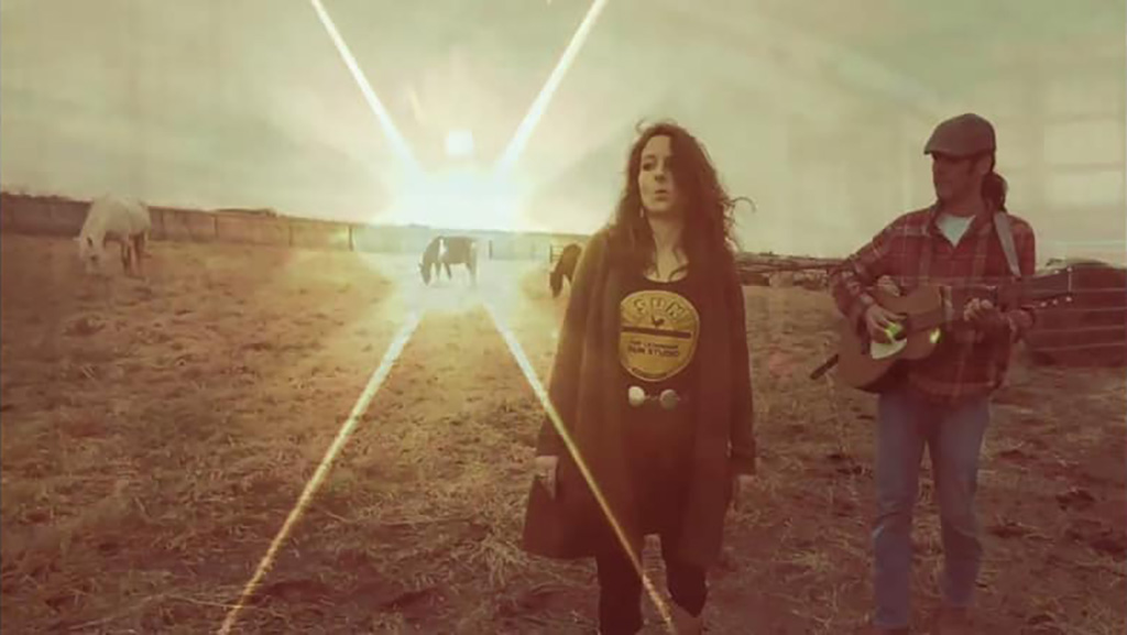 The two members of Black Eyed Lillies in a field with the sun just over the horizon behind them. The male is holding a guitar and looking off to the write. The singer is turned to the left.