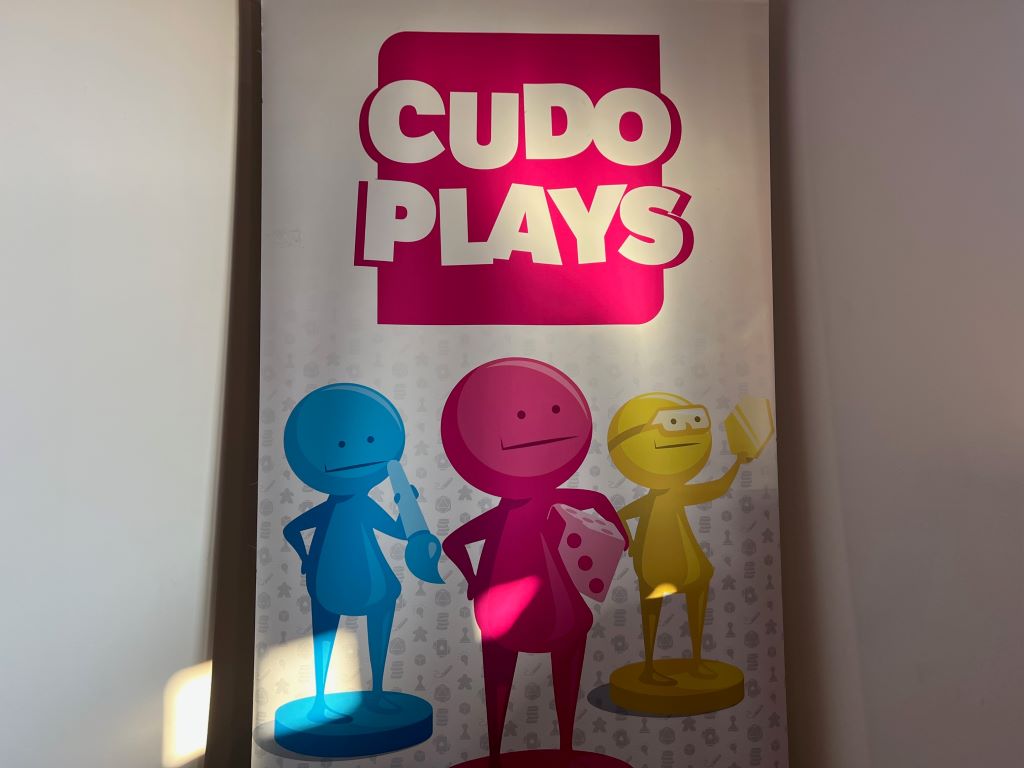 A white vertical banner with a pink square and the words CUDO PLAYS in white block letters at the top. Underneath there are three figures, one blue, one pink, and one yellow.
