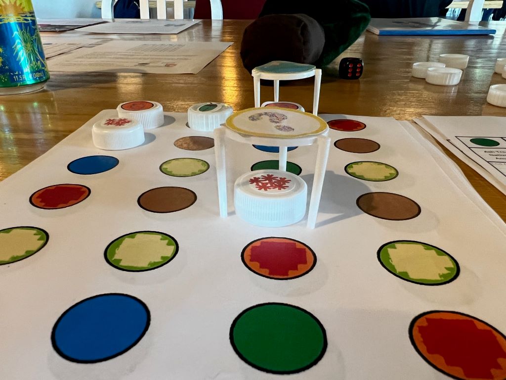 A white piece of paper covered in rows and columns of different colored dots. There are white bottle caps covering some of the dots, and two markers made from plastic pizza box stands on the paper.