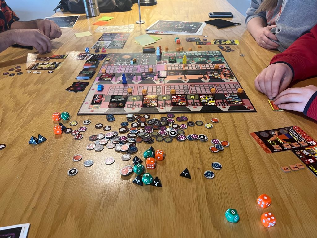 Close up of a board games with colorful squares. Surrounding the game are piles of cards, dice, and small colorful discs. There are three people's hands visible at the table.