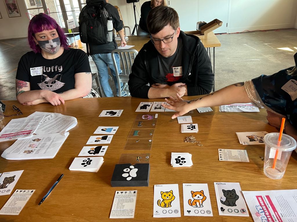 The setup of a card game with pictures of cats, clear cards with outfit pieces on them, cards with cartoon images of people, and small fish shaped pieces. There are three people around the table, one watching, one playing the game, and one gesturing over the set up.