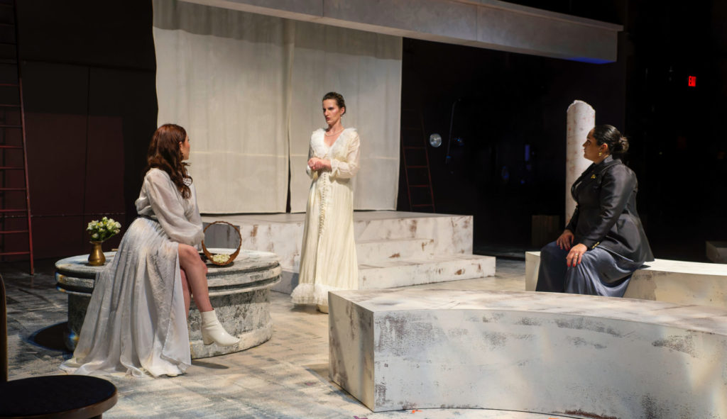 A scene from Coriolanus. Three women are on the set. One is seated to the left wearing a long, light gray gown. One woman stands in the middle facing gray gown woman. She wears a long white gown. On the right is another seated woman wearing a black blazer. 