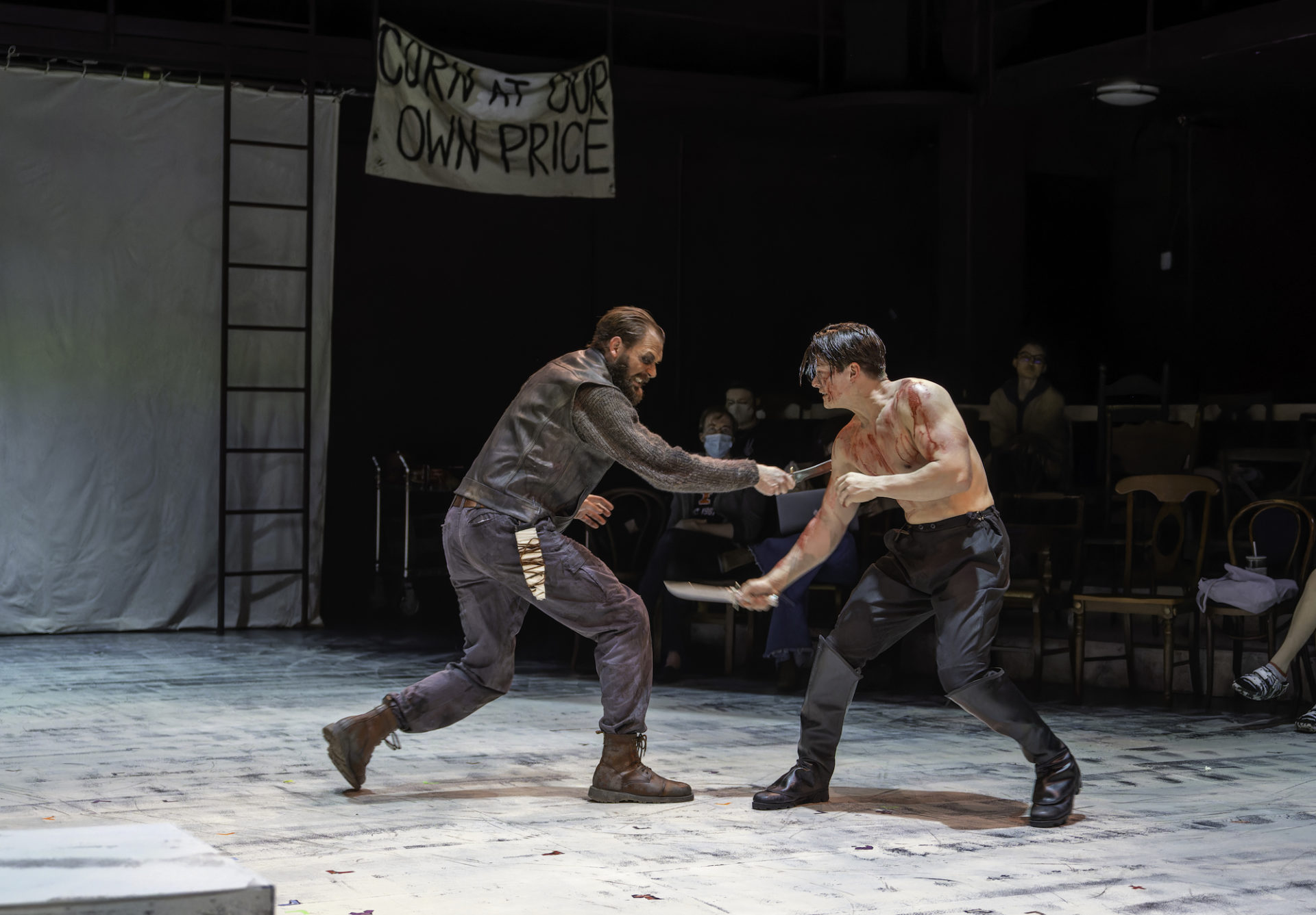 Two men engage in a fight scene. The man on the left is fully clothed in pants, boots, and a jacket. He is attempting to stab the man on the right who is also holding a knife. The man on the right appears to be covered in blood and is shirtless.