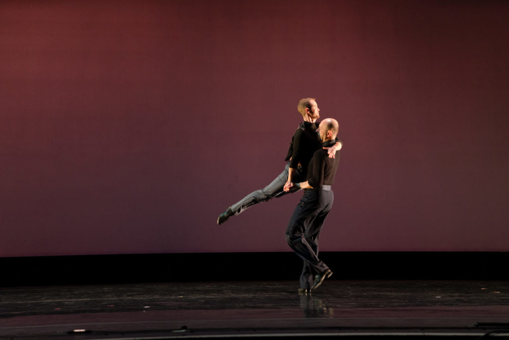 Alex Tecza and Kato Lindholm perform a tango on stage. Tecza is holding Lindholm in a traditional ballroom pose in the air, with his legs extended.
