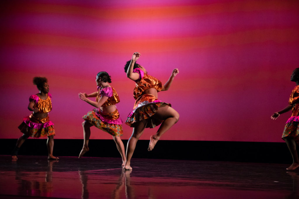 A group of female dancers wearing crop tops and matching skirts in orange, pink, and African print, perform "Women Gather" against a pink background on stage. 