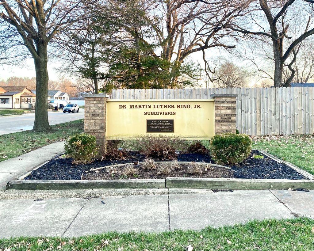 A yellow sign with two brick columns sits in a square area landscaped with mulch and small green bushes. It says Dr. Martin Luther King Jr. Subdivision. Behind the sign is a wooden fence and trees. 