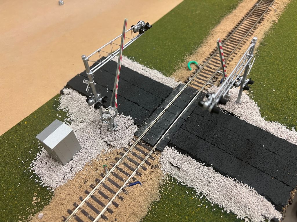 Close up of a model railroad crossing, with green turf, a track running through the center, and railroad signals on either side.