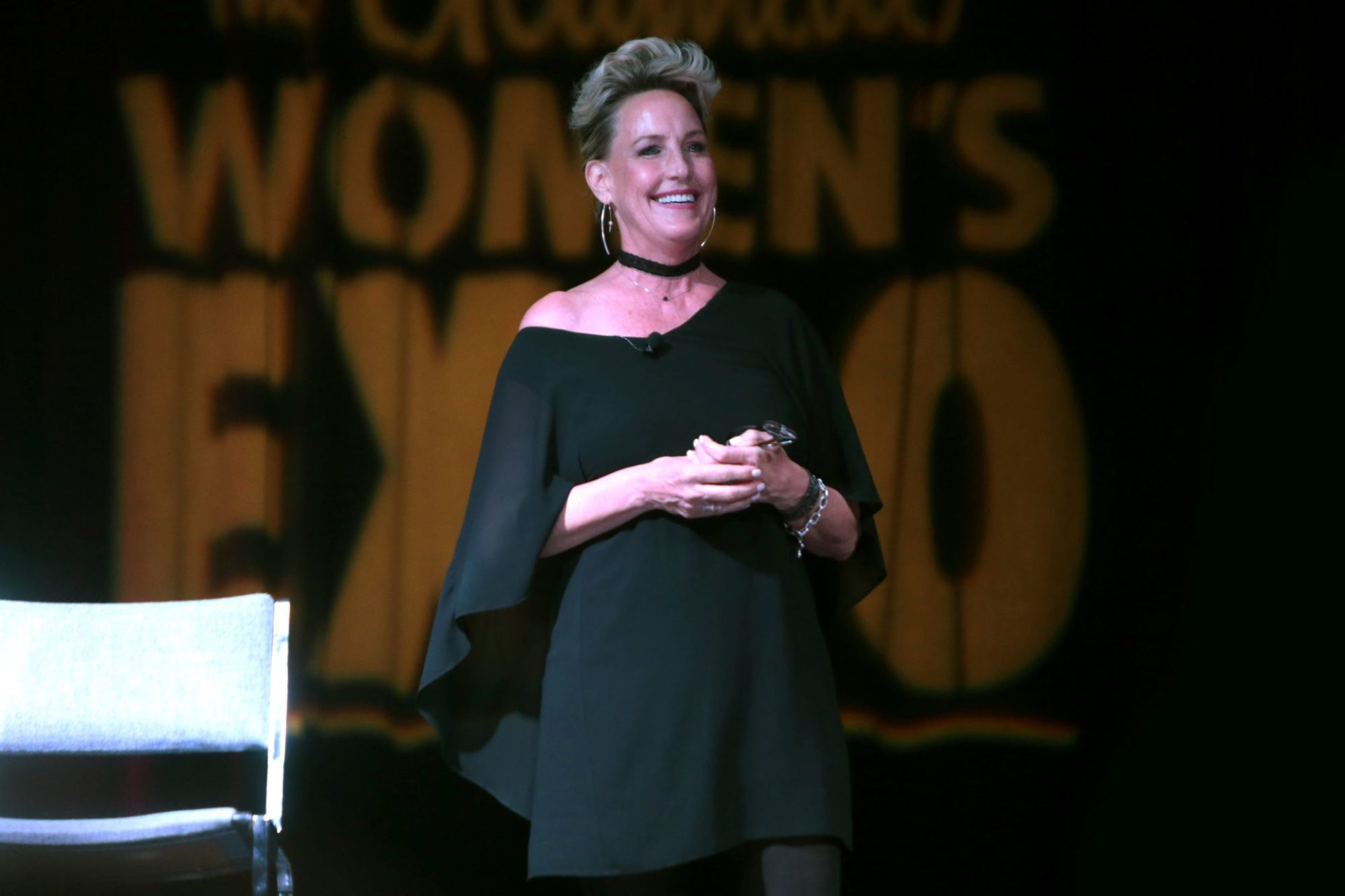 A white woman with blond hair is standing on a stage. She is wearing a black dress, and smiling out at the audience.