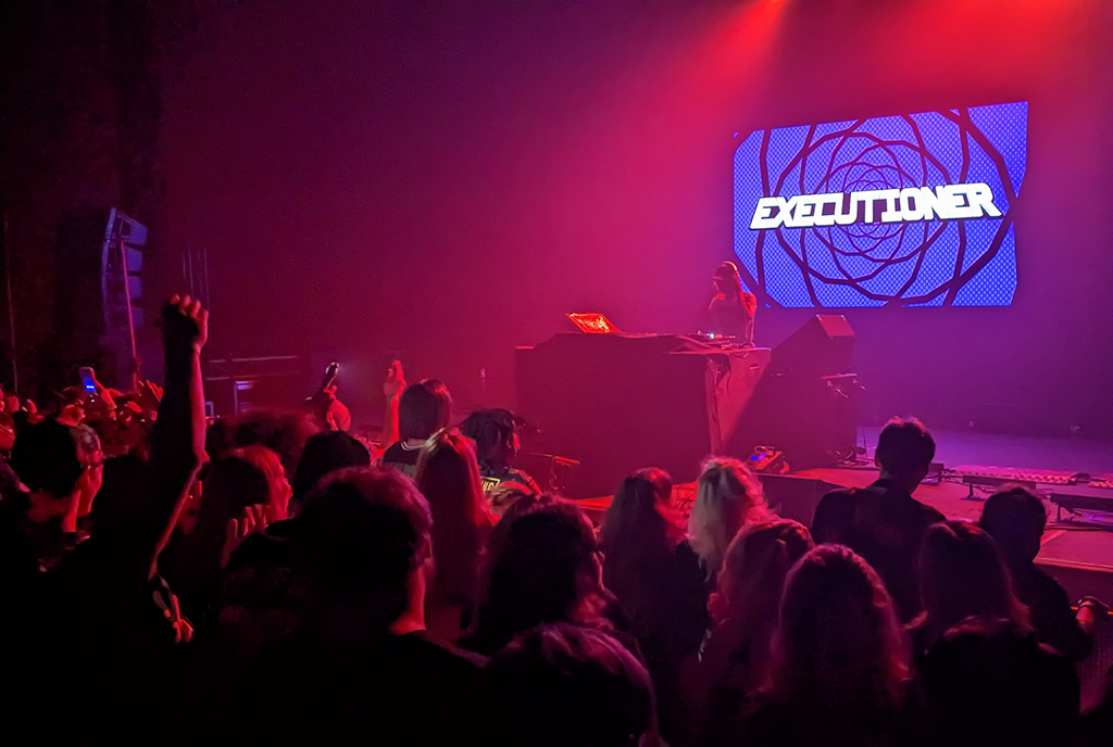 Front rows and stage at an EDM show. Red light fills the stage. Executioner is playing his music on the decks onstage, and a video screen with "Executioner" on it is behind him.