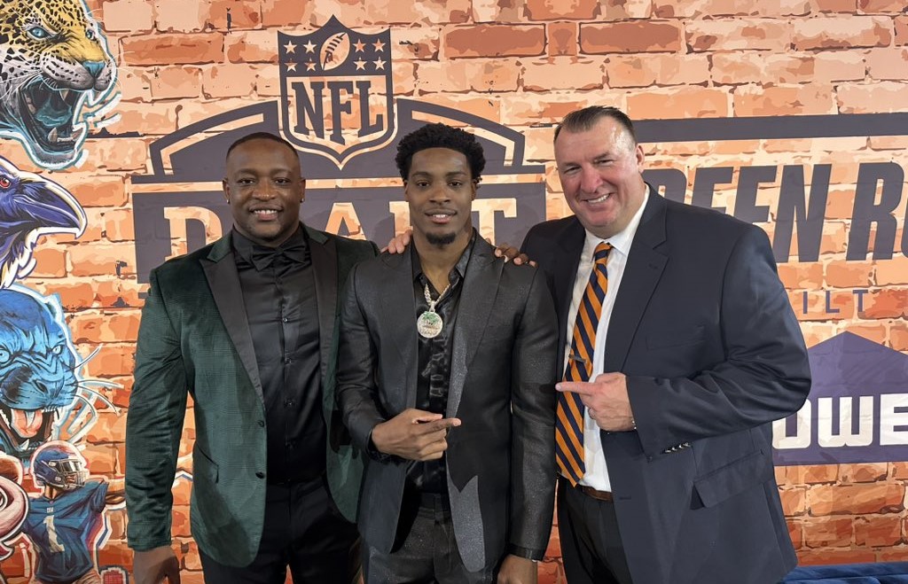 Two Black men and one white man are standing together in front of a backdrop with an NFL logo.