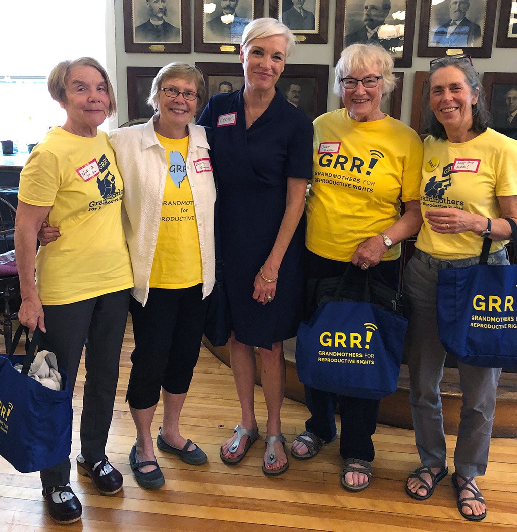 A group of four older women in bright yellow t-shirts posting with a woman in a dark blue dress with short gray hair. 