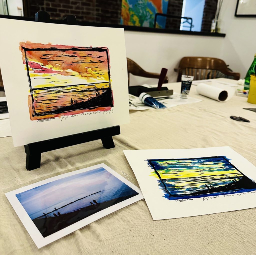 An artists worktable: a photograph of the beach and ocean at night lays on the table with two mixed media recreation paintings next to it