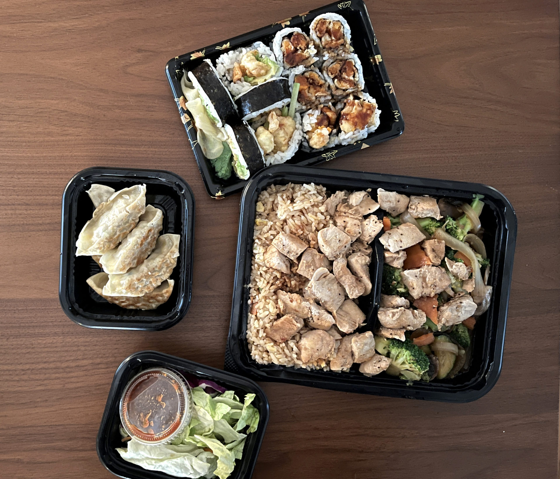 Four take out containers with food from Nori Sushi. Clockwise, from top: chicken tempura and sweet potato tempura sushi, hibachi chicken, side salad, gyoza.