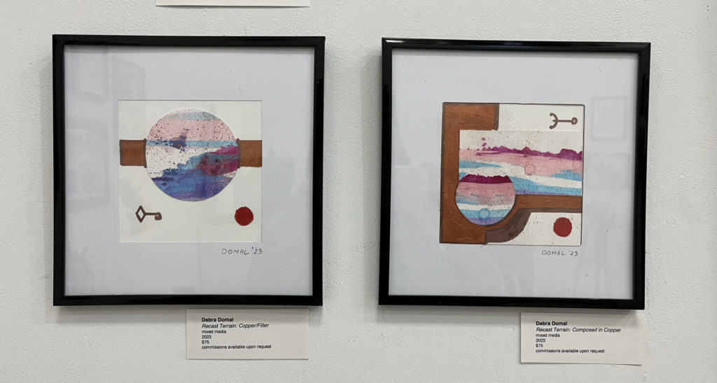 Two small, framed works on paper. On the left, a circle with pinks and blues overlaid on a copper line. Below is a key and a red dot. On the right, a similar circle overlaid atop a vertical copper rectangle and a horizontal colorful rectangle. Above is a symbol and below the rectangle is a red dot.