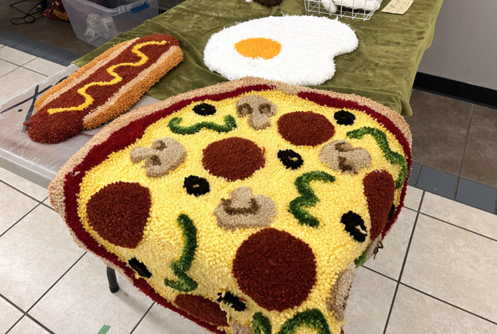 A display table of the textile work by High in Fiber Rugs. On the table are three large pieces meant to resemble: a hot dog, an egg, and a pizza with pepperoni, mushrooms, olives, and green peppers.