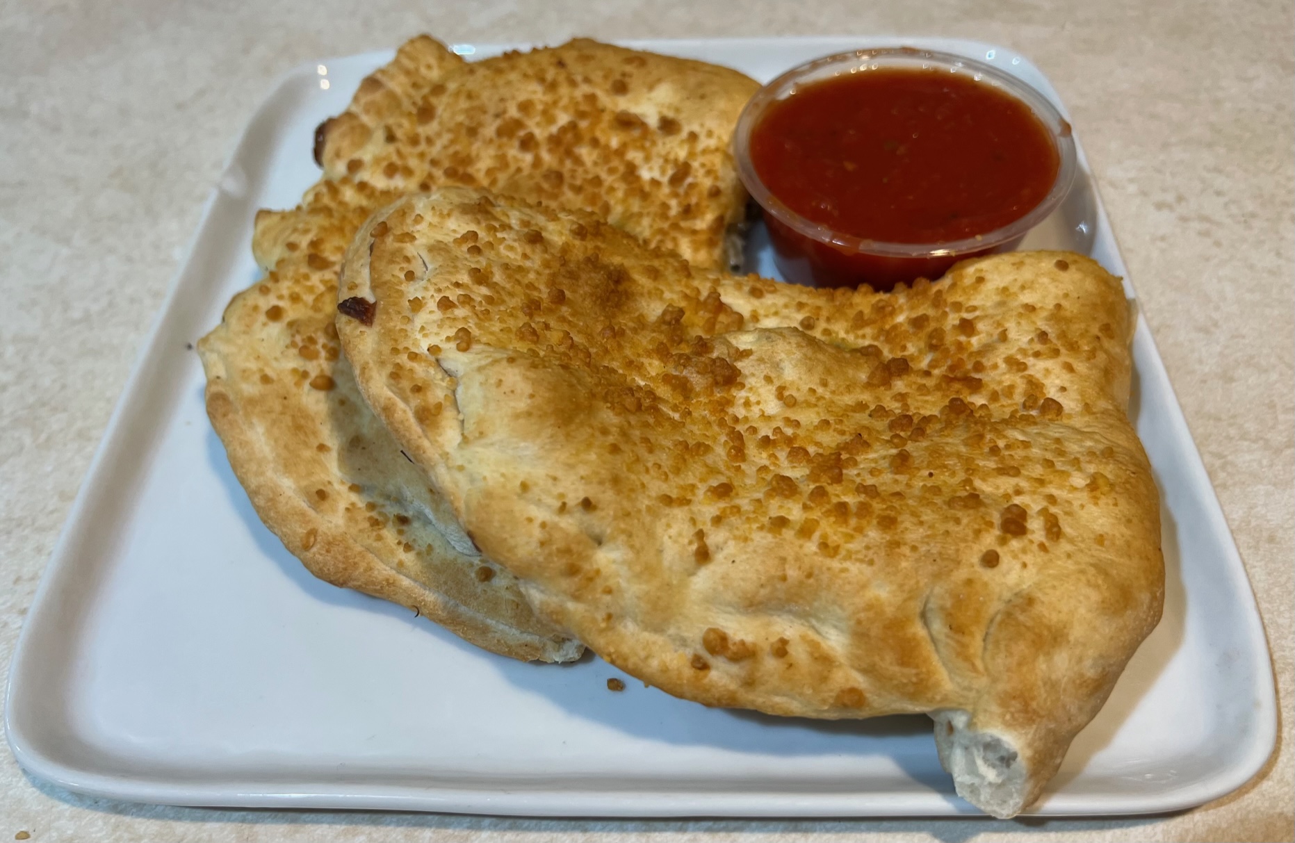 Two halves of a calzone sit on a square white plate. There is a small cup of marinara sauce next to them.