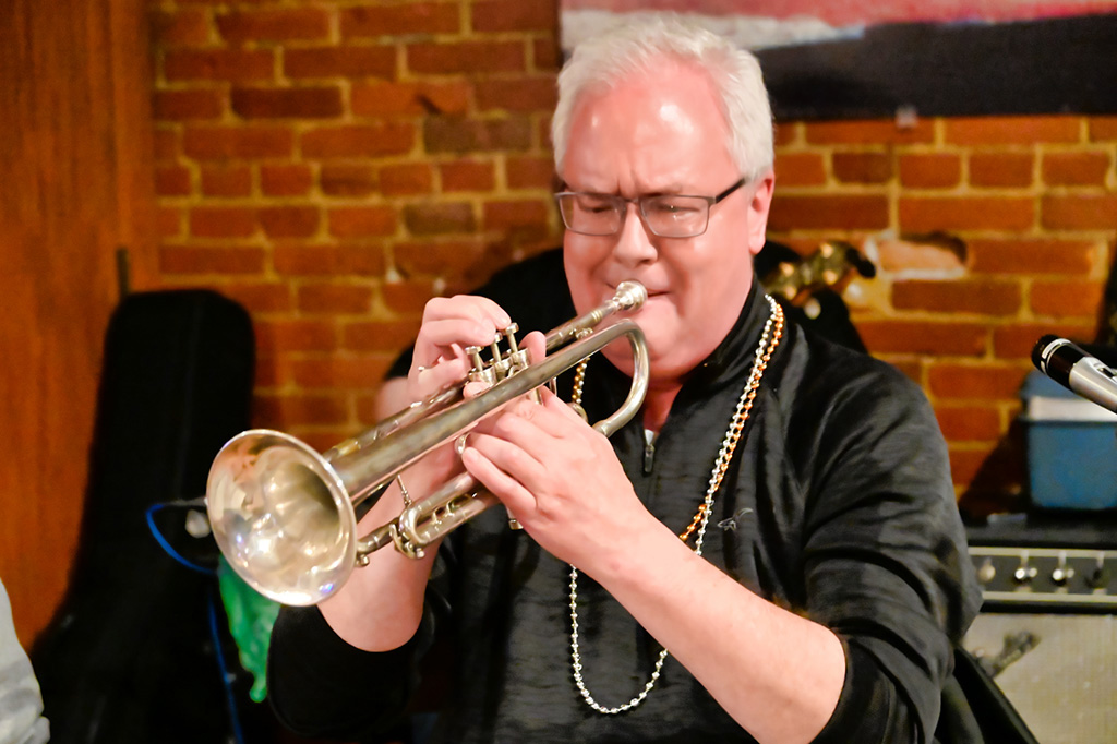 Jeff Hegelsen wearing a black shirt, mardi gras beads and playing the trumpet.