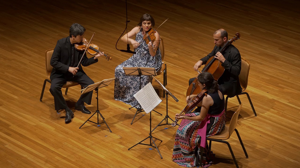 4 members of Jupiter String Quartet playing on a stage with a hardwood floor. All musicians are seated. There are 3 violin players and a cello player.
