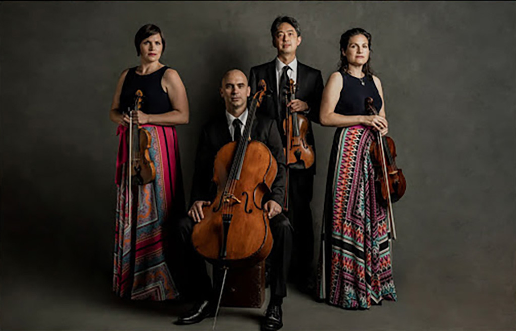4 members of the Jupiter Quartet against a dark background. The 3 violin players are standing and the cello player is sitting.