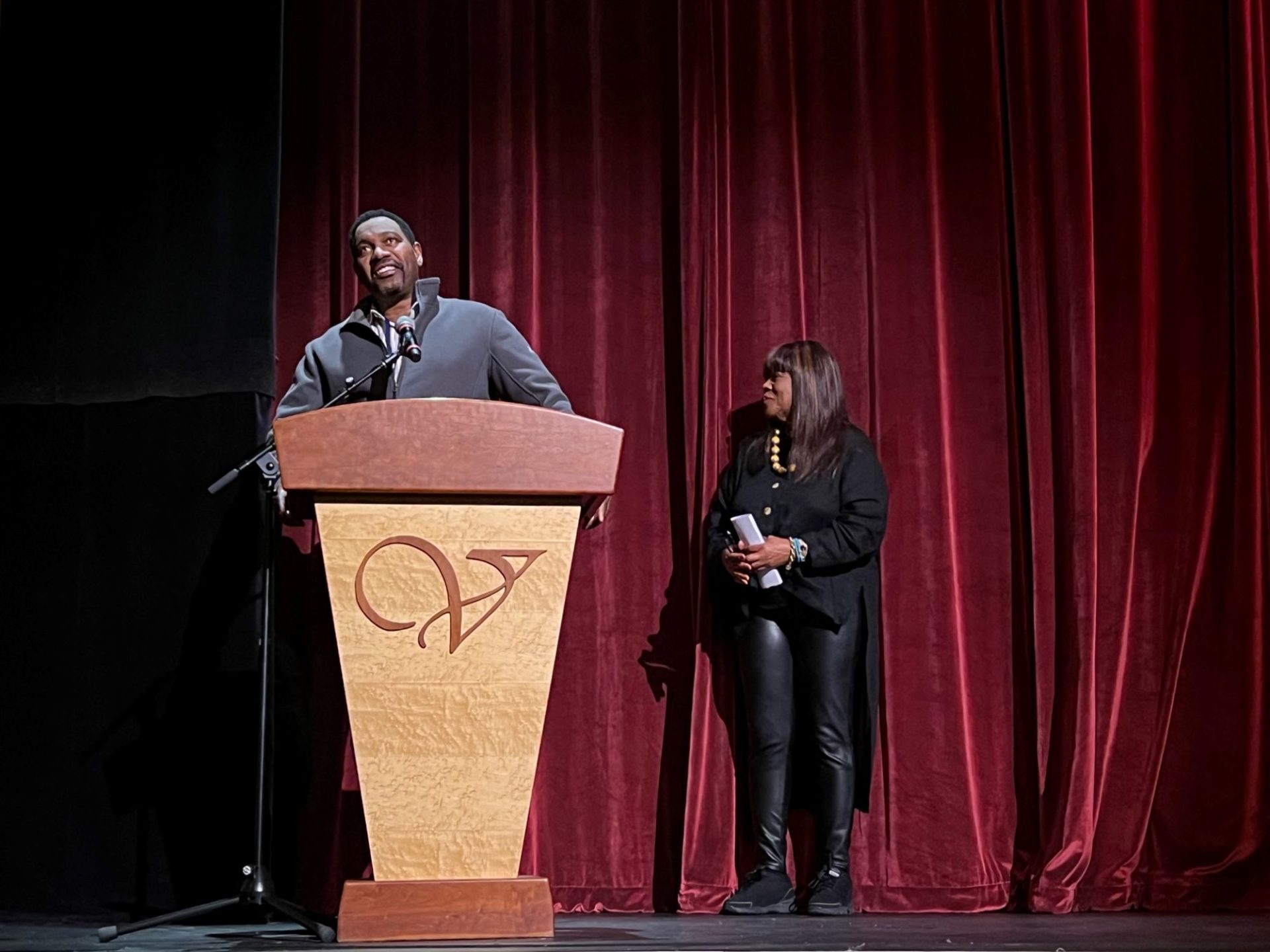 A Black man in a gray sweater is standing behind a wooden podium with a microphone. A Black woman with long brown hair and a black jacket and pants is standing near him, looking to the side.