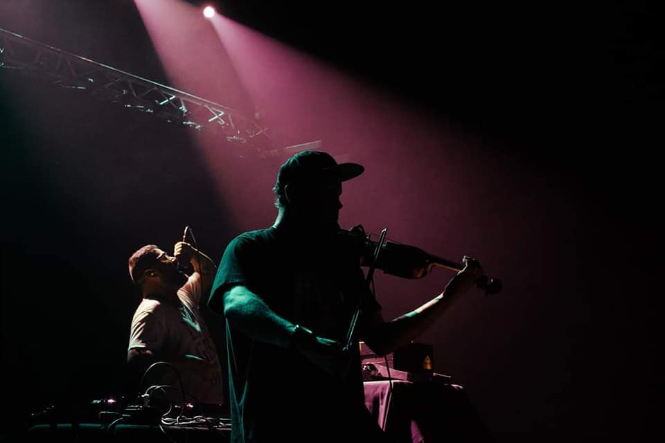 Dimly lit stage with a shadow of a person playing violin towards the front and a DJ singing into a mic in the background.