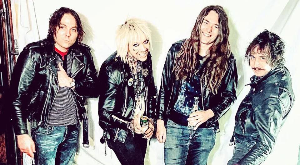 4 leather-jacket-clad members of Mid Nite Leg in various poses standing in front of a white wall.