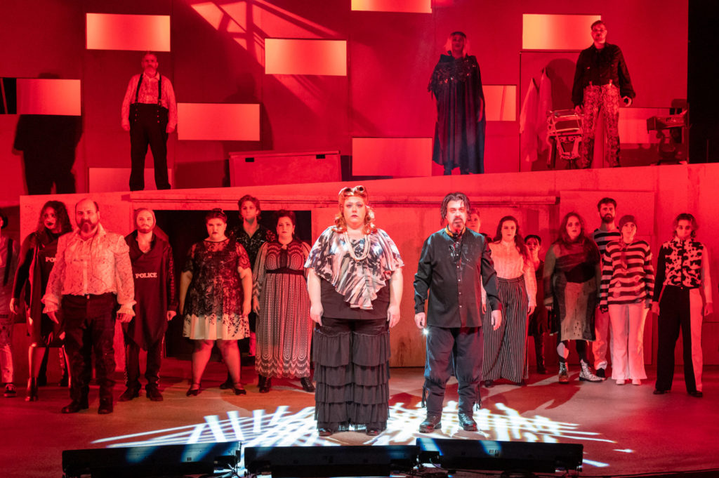 The entire cast of Sweeney Todd stands still in red lighting, somber faces, staring at the audience. Matt Hester and Nicole Morgan who play Sweeney Todd and Mrs. Lovett are standing front and center with the rest of the cast behind. 