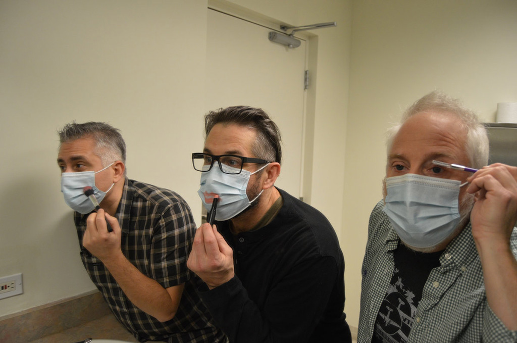 Three men with surgical masks on, looking into a mirror and trying to apply makeup to their faces and the masks.