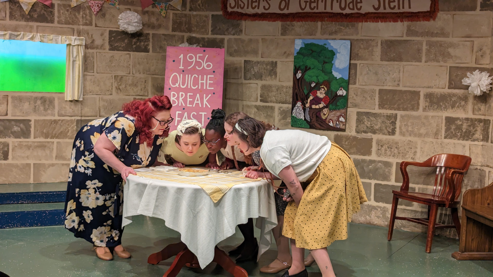 Photo of the set with cast, 5 Lesbians Eating a Quiche at the Station Theatre. Four white women and one Black woman are leaning over a small round table smelling a quiche. They are dressed as women from the 1950s. L-R - Heather Smith-Holley, Ellen Magee, Tiphaine Kouadou, Zoë Dunn, Erin Roux
