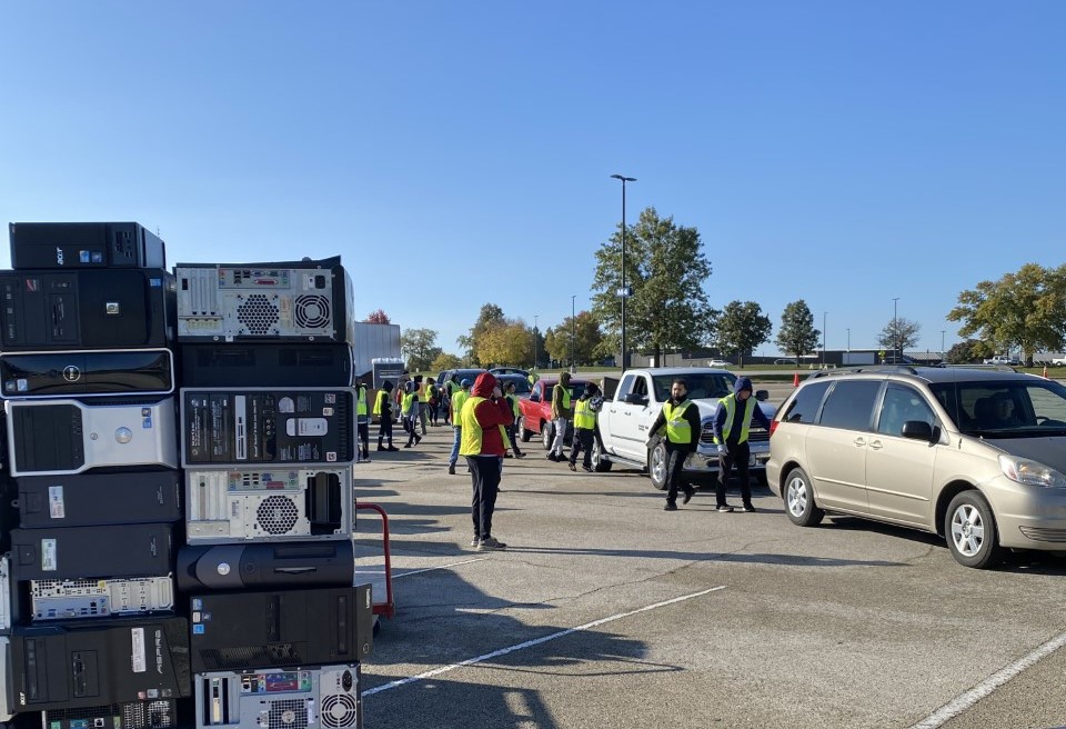 A line of cars in a parking lot, with several people in bright yellow vests approaching the cars. In the foreground is a stack of electronic consoles.