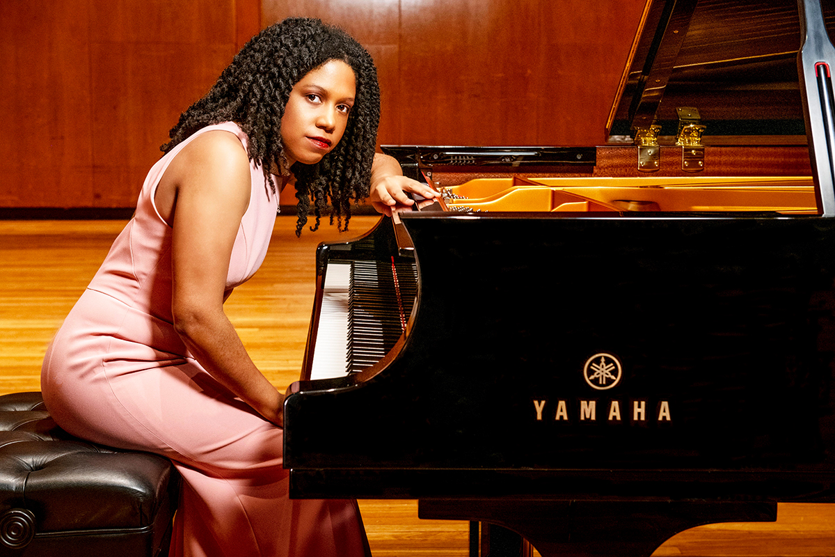 The image captures a scene where a person with curly hair is seated at a grand piano. The individual is wearing a fitted, sleeveless pink dress that reaches down to their knees. The grand piano, glossy and black, has its lid open, revealing its intricate internal structure and strings. The setting is a room with polished wooden floors. Two small glass bottles are placed on top of the piano. The overall ambiance is elegant and serene.