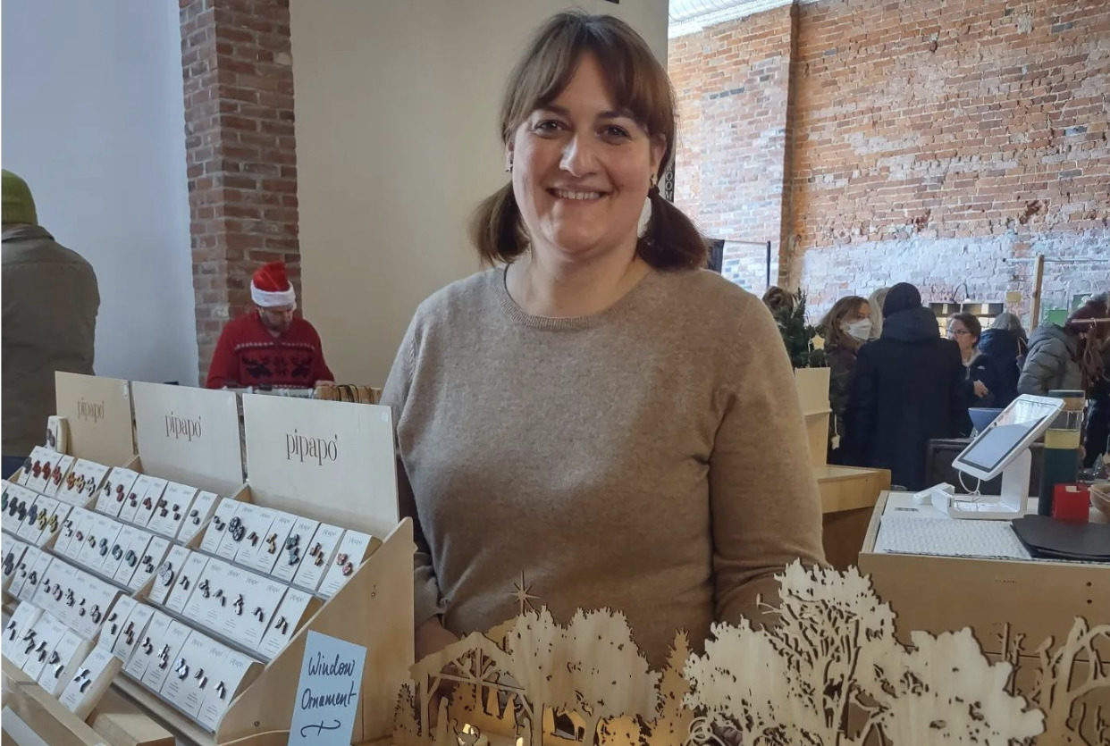 Anna Gutsch, a white woman, is wearing a beige sweater and standing behind a display of birch jewelry and home decor.
