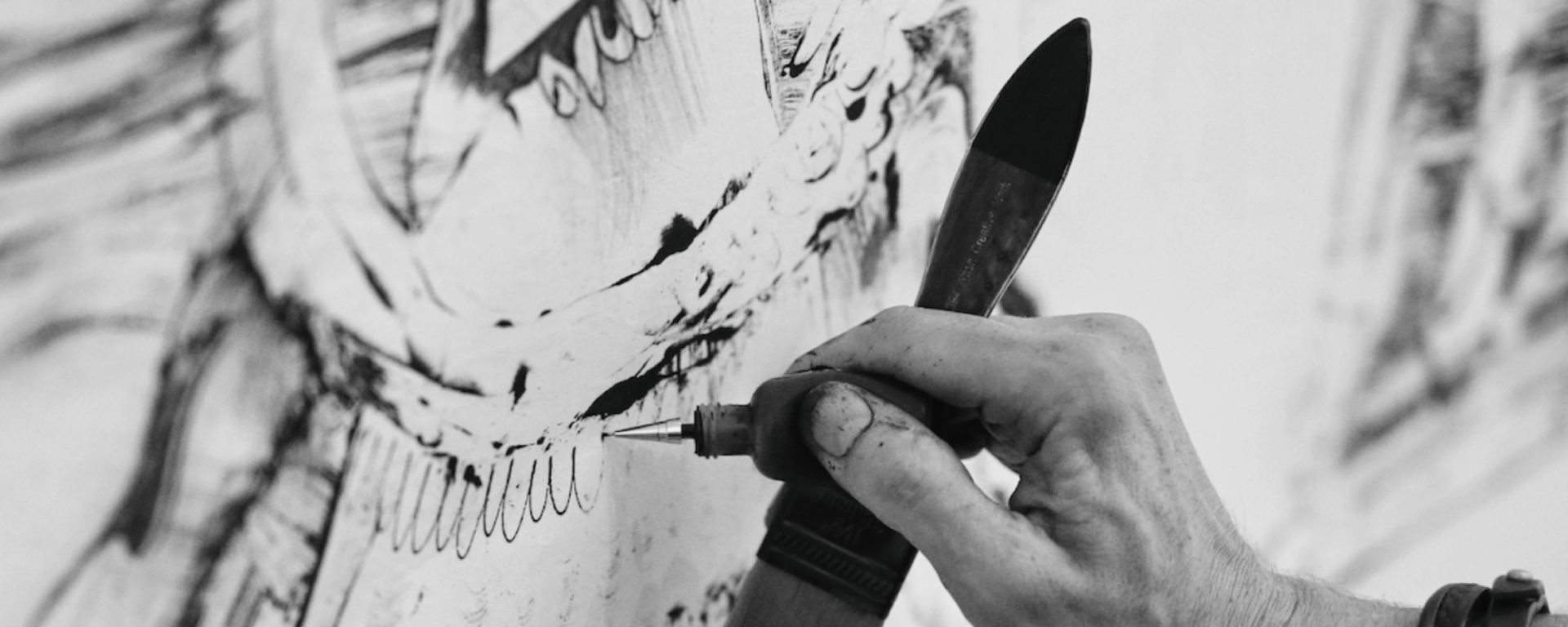 Close up of artist Kevork Mourad making a drawing. The image is cropped around his hand holding a drawing tool and brush as he makes marks on a white piece of paper.