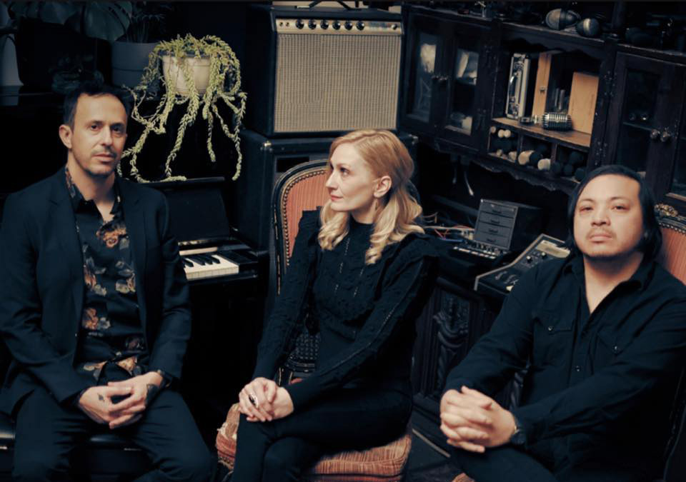 3 members of Spotlights sitting in chairs in a small corner of a recording studio. All are dressed in black. The male band members are looking at the camera, but the female band member sitting in the middle is looking off to the left.