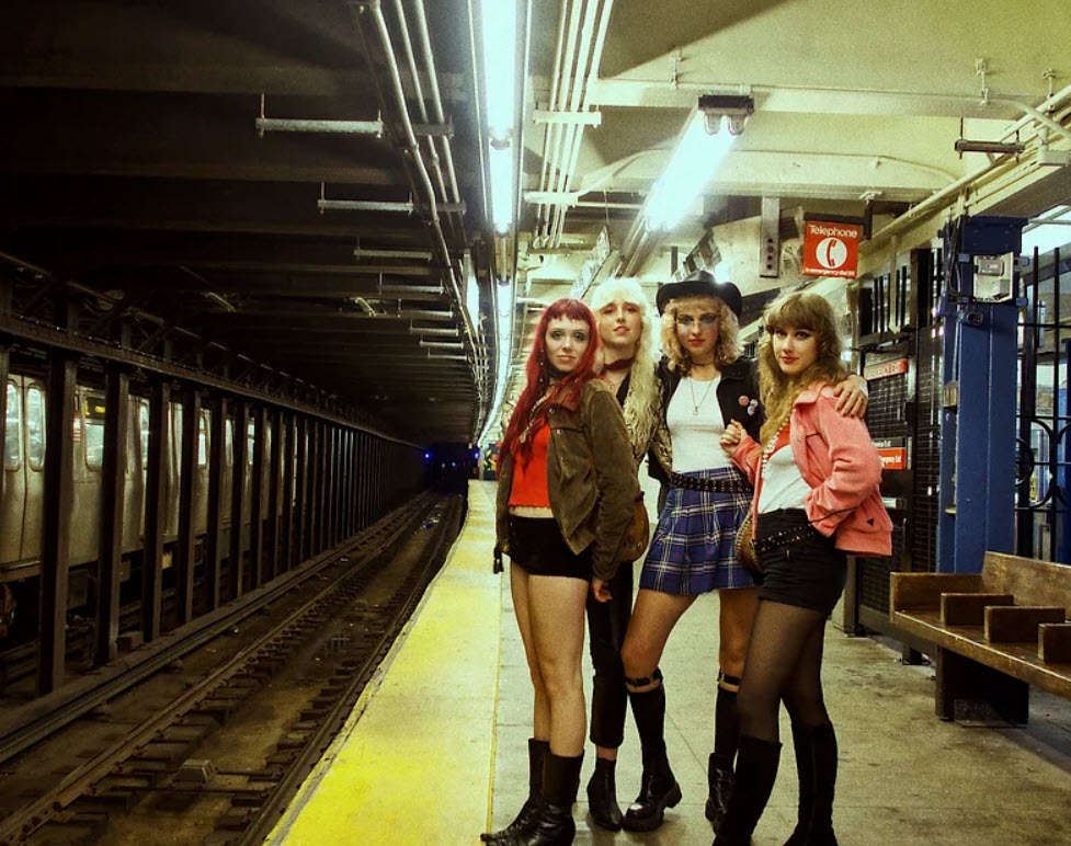The four members of the band The Knee Hi's on a subway platform, huddled together and looking at the camera.