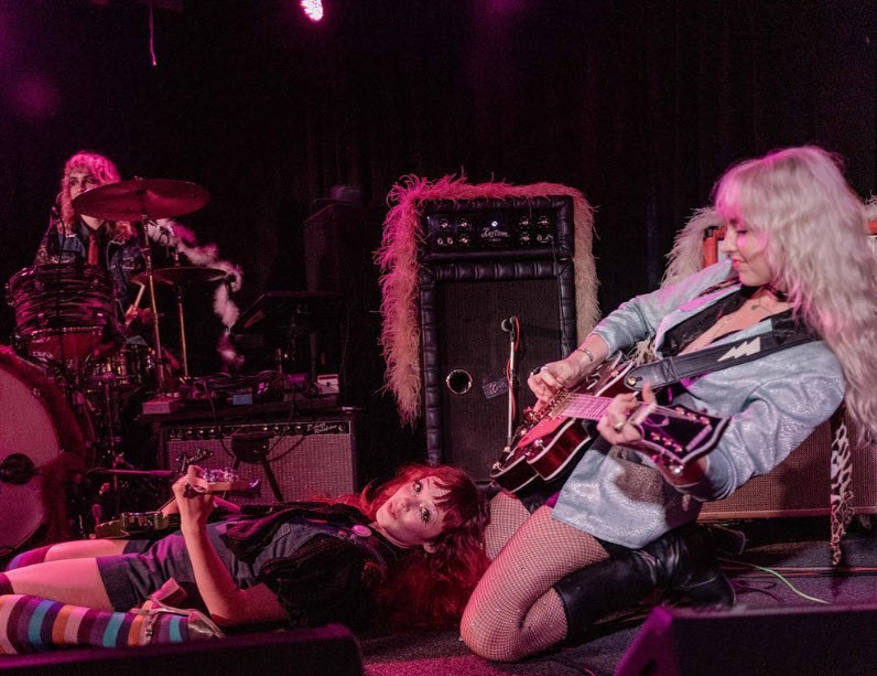 3 members of The Knee Hi's onstage. Chris is behind the drums in the back. Bassist Dev is laying on her back with her feet to the side. To the right is guitarsit Ash kneeling while bending back all while strumming her guitar.