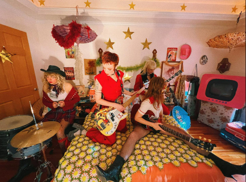 The four member of The Knee Hi's playing their instruments while crammed into someone's colorful bedroom. The drummer is to the left of the bed and the rest of the members are sitting or kneeling on the bed. No one is looking at the camera.